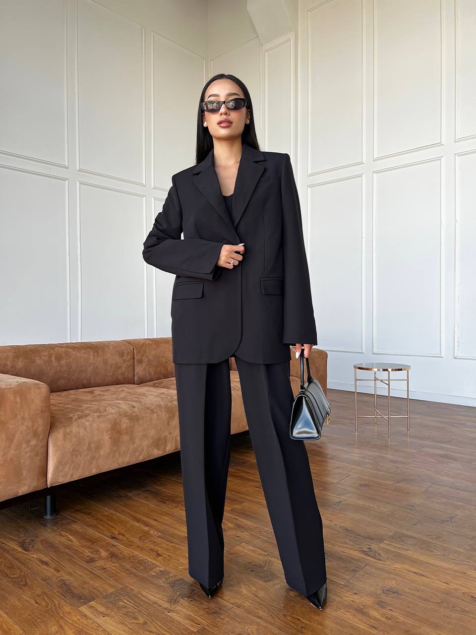 Woman in a black suit standing in a room with a brown couch and white paneled walls.