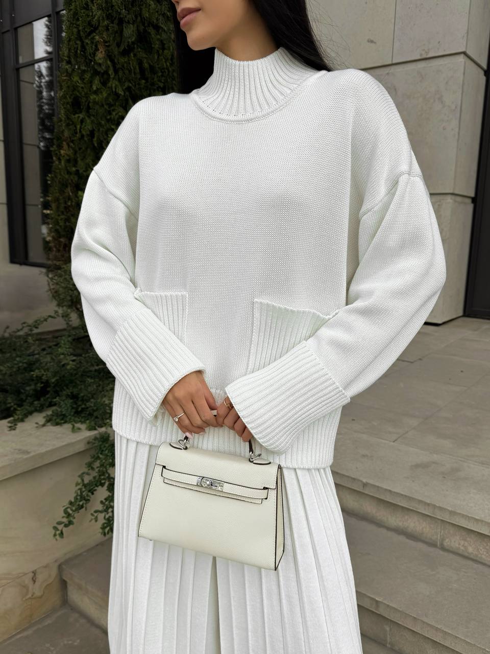 Woman wearing a white knit sweater and pleated skirt holding a matching handbag.