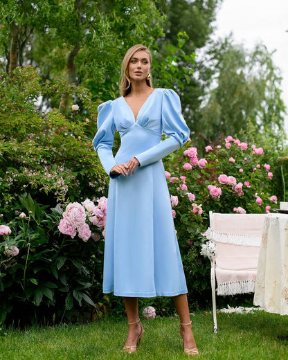 Woman in a light blue dress standing outdoors with flowers and greenery in the background