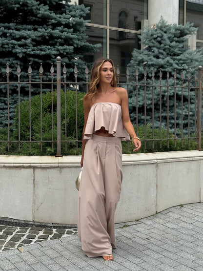 Woman in a beige outfit standing outdoors with greenery and a building in the background
