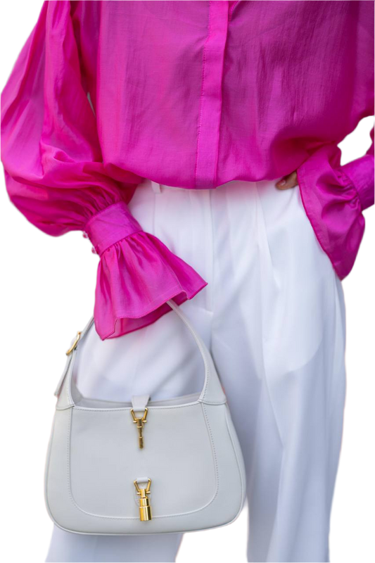 Person wearing a bright pink blouse and white pants holding a light gray handbag with gold accents.