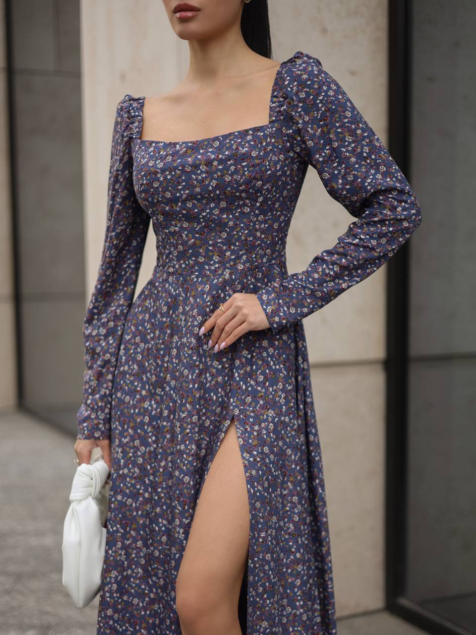 Woman wearing a floral dress with a high slit, holding a white clutch against a neutral background.