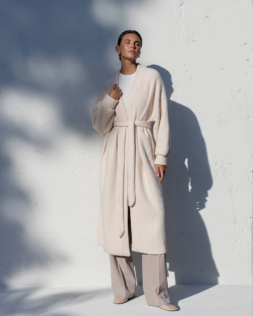 Person wearing a long beige coat against a white wall