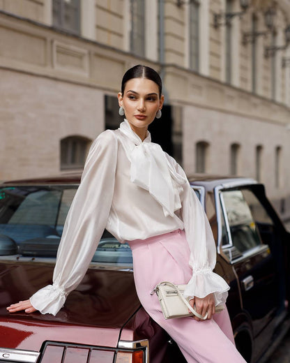 Woman in a white blouse and pink pants standing next to a vintage car with a building in the background