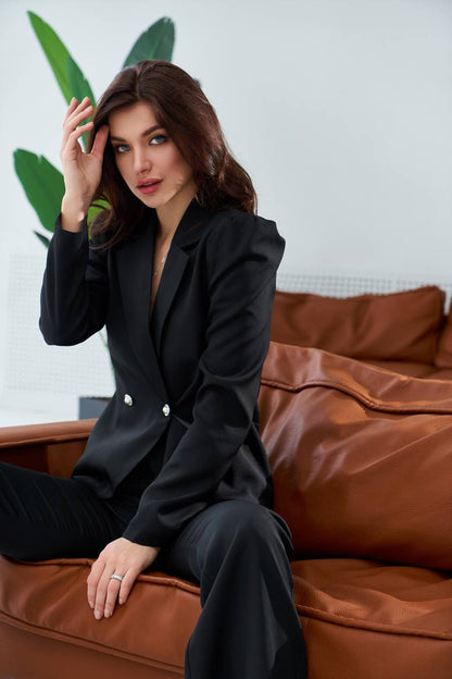 Woman in a black suit sitting on a brown leather couch with a white wall and green leaves in the background