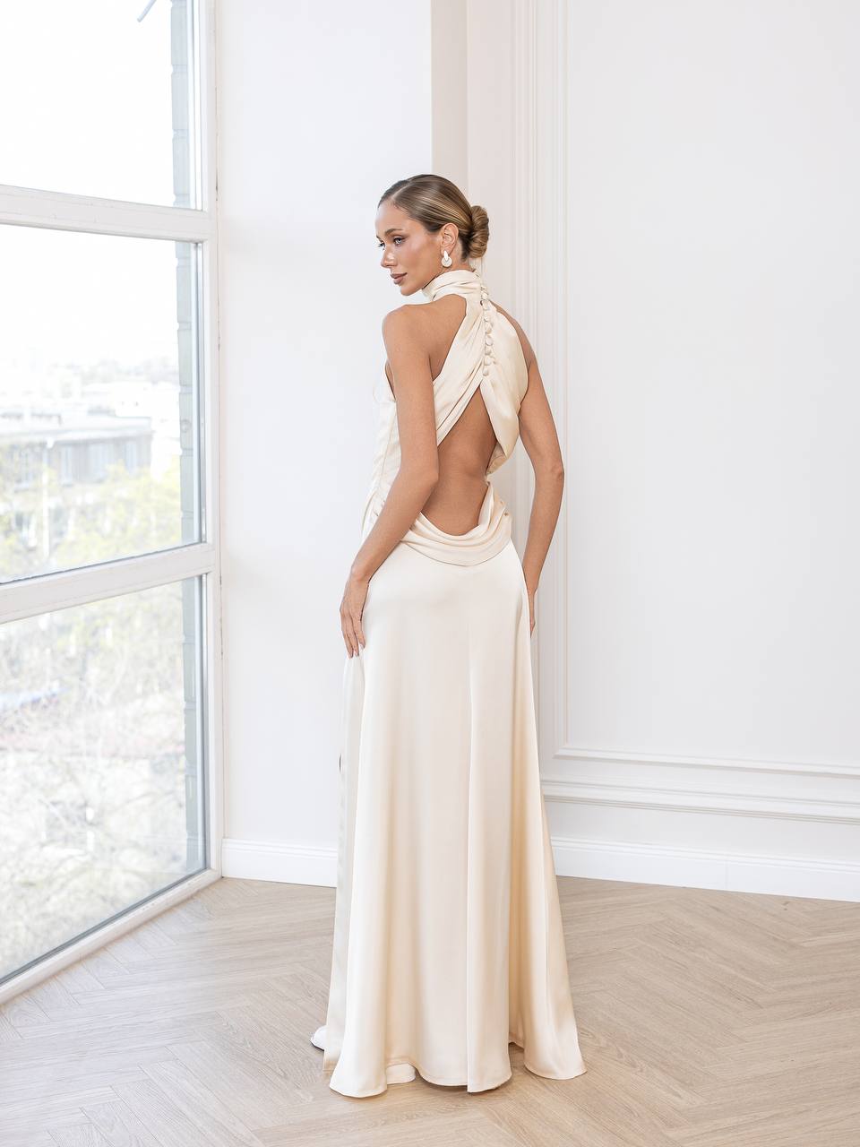 Woman in a long, elegant white dress standing in a bright room with large windows.
