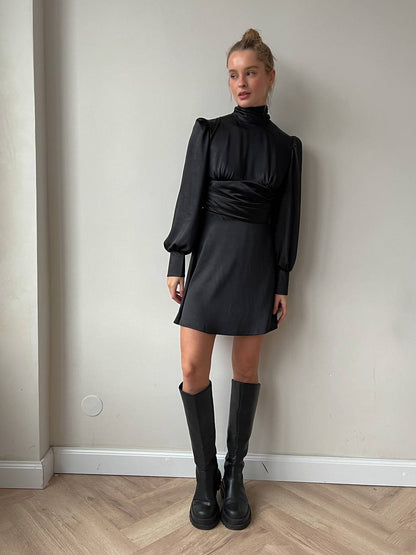 Person wearing a black dress and boots against a plain wall.