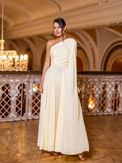 Woman in a light-colored one-shoulder dress standing in an elegant indoor setting with chandeliers.