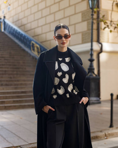 Woman wearing a black coat and patterned sweater standing on a street.