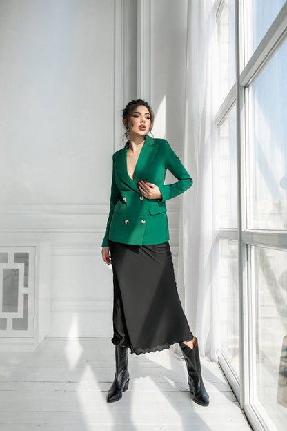 Woman wearing a green blazer and black skirt standing in a bright room with large windows.