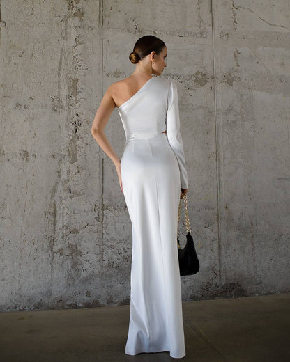 Woman in a white one-shoulder dress holding a black handbag against a textured wall.