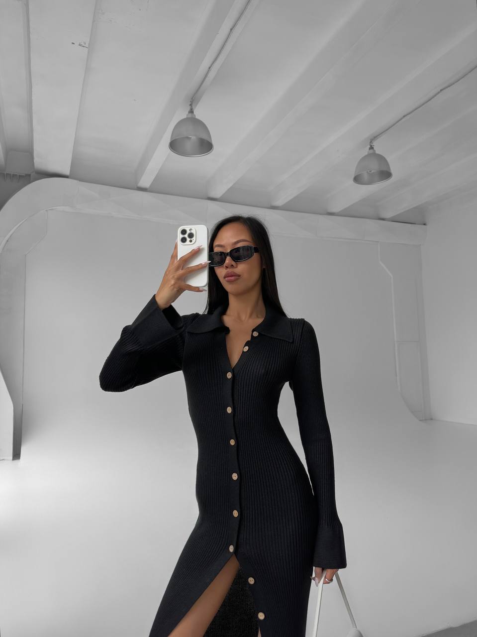 Person wearing a black dress with a slit, standing in a minimalistic room.