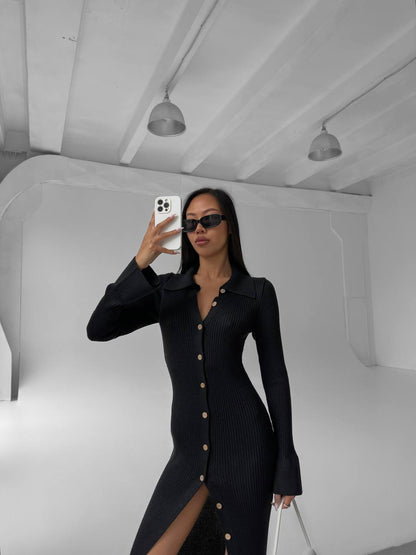 Person wearing a black dress with a slit, standing in a minimalistic room.