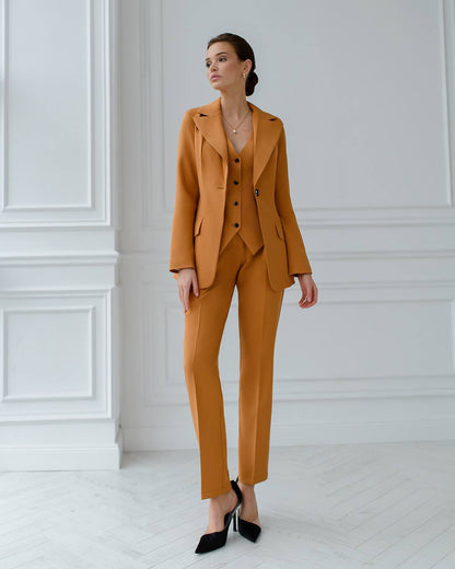 Woman wearing a mustard yellow suit standing against a white paneled wall.