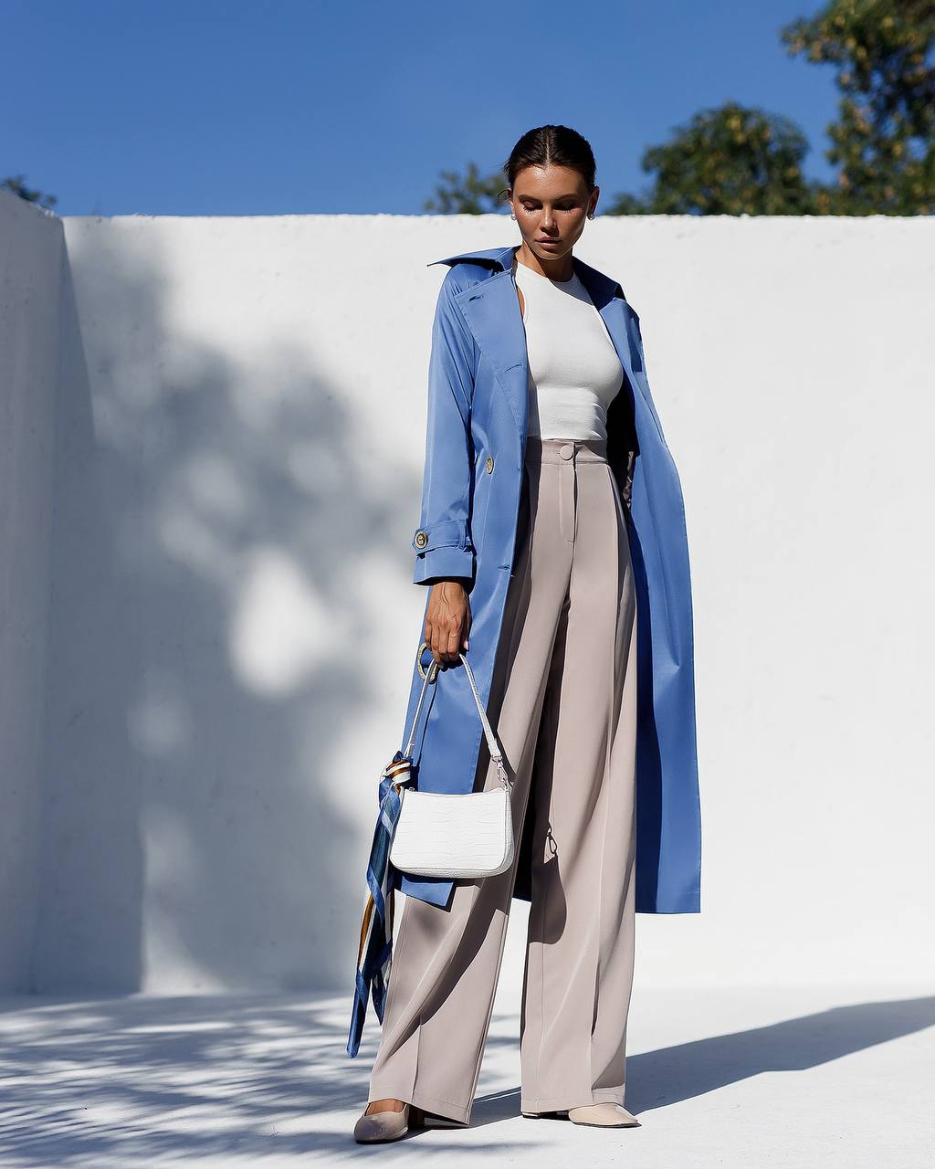 Woman wearing a blue coat and beige pants standing against a white wall with a blue sky.