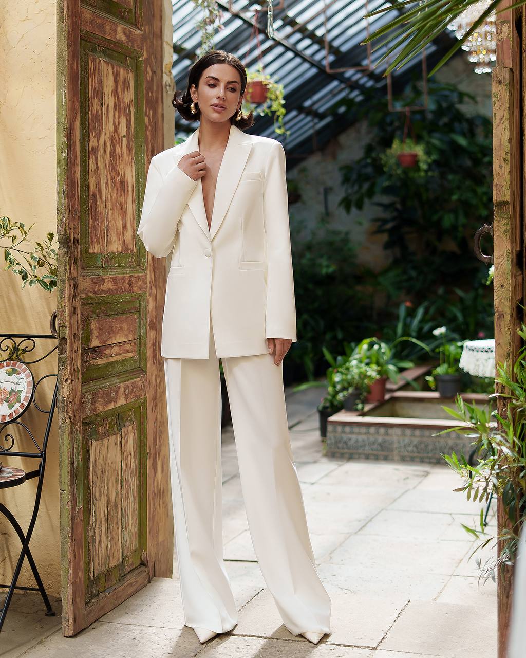 Woman in a white suit standing in an outdoor setting with plants and a wooden door.