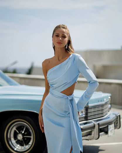 Woman in a light blue dress standing next to a classic car with a clear sky background