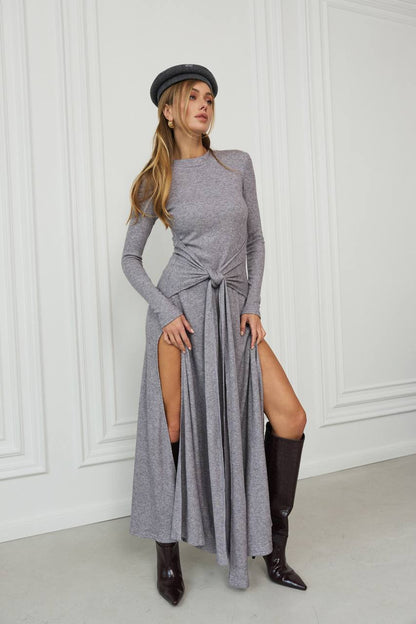 Woman wearing a long gray dress with a slit, black knee-high boots, and a black beret against a white paneled wall.