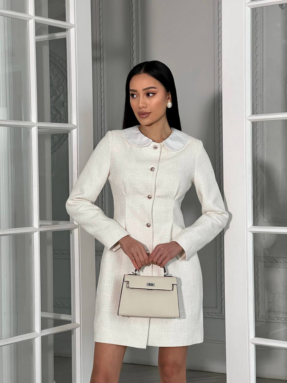 Woman in a white dress holding a beige handbag in front of a glass door.