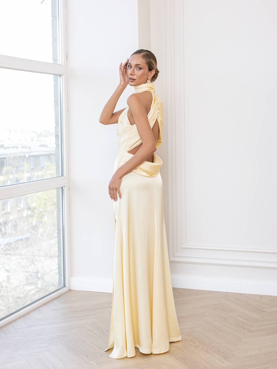 Woman in a light yellow dress standing in a bright room with large windows.