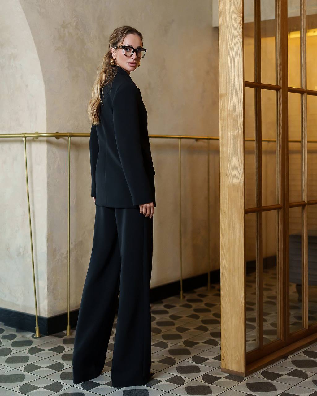 Woman in a black outfit standing in an elegant interior setting with wooden panels and tiled floor.