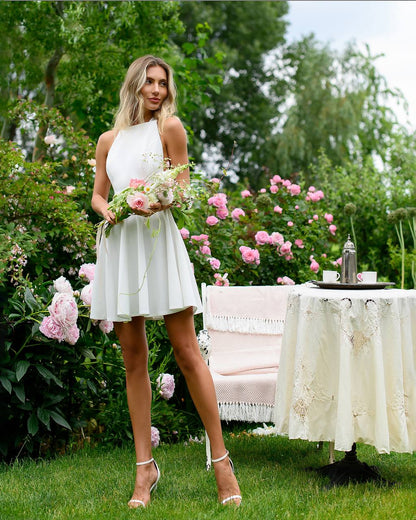 Woman in a white dress holding flowers in a garden setting with a table and chairs.