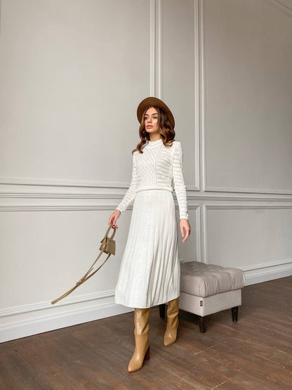Woman in a white outfit with brown boots and hat standing against a neutral wall.