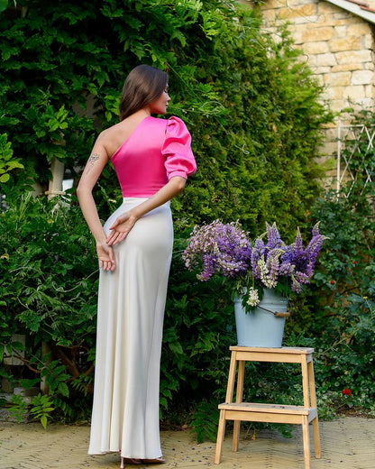 Woman in a pink top and white pants standing next to a wooden stool with flowers in a garden setting.