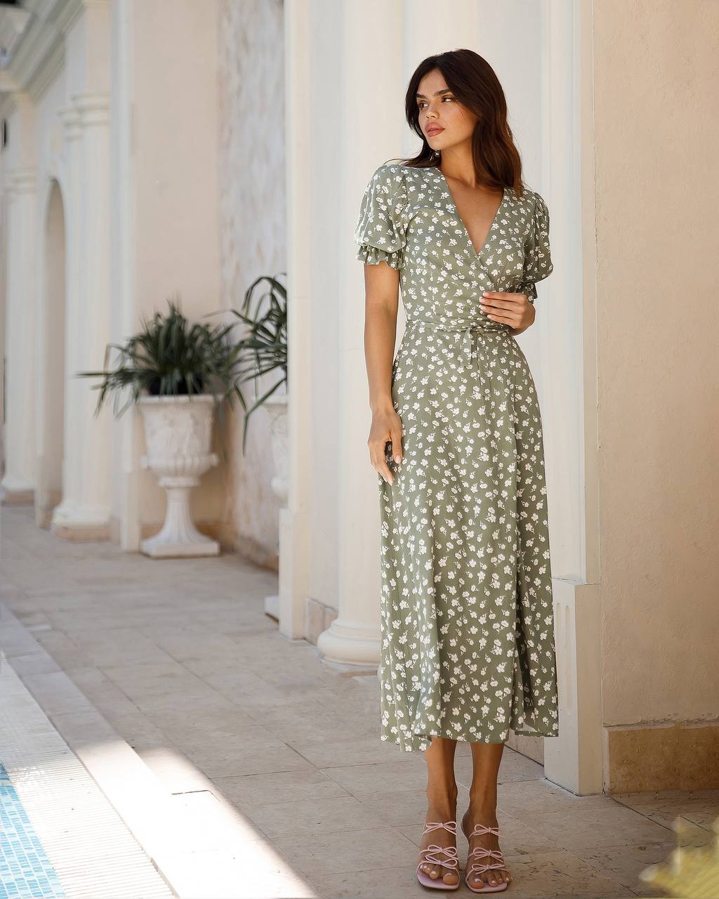 Woman in a green floral dress standing in a sunlit outdoor setting with columns and plants.