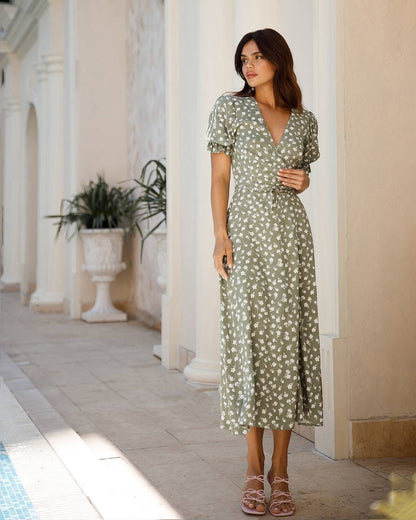 Woman in a green floral dress standing in a sunlit outdoor setting with columns and plants.