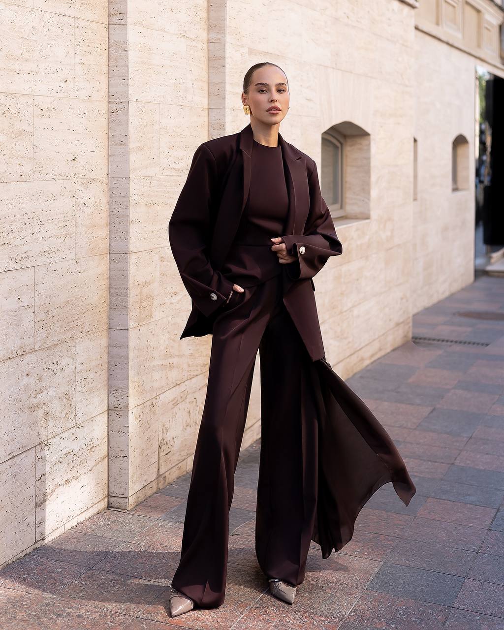 Person wearing a dark brown outfit standing against a light stone wall.