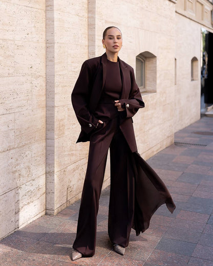 Person wearing a dark brown outfit standing against a light stone wall.