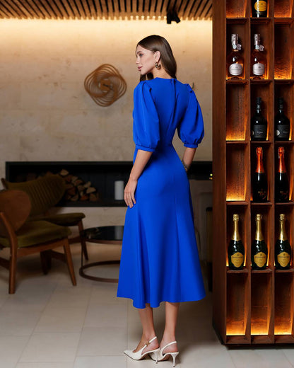 Woman in a blue dress standing in a stylish room with a wooden shelf of bottles.