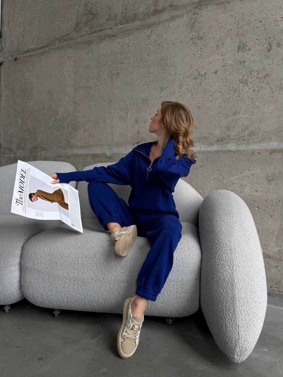 Person in blue outfit reading a newspaper on a modern gray sofa against a concrete wall.