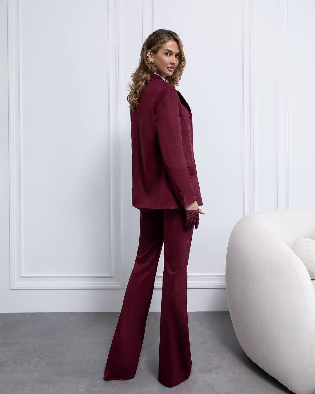 Woman wearing a burgundy suit standing in a room with white walls and a white chair.