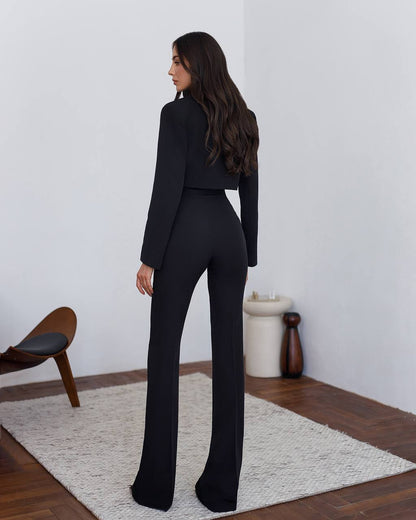 Woman in a black suit standing in a minimalistic room with a white wall and wooden floor.