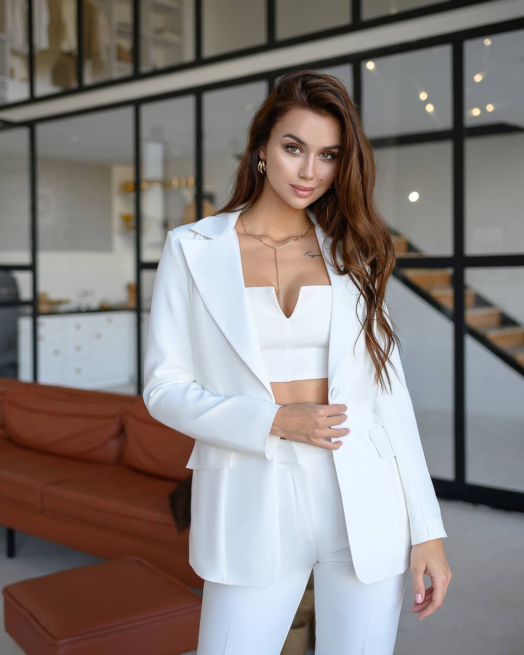 Woman in a white outfit standing in a modern indoor setting with glass walls and brown furniture.