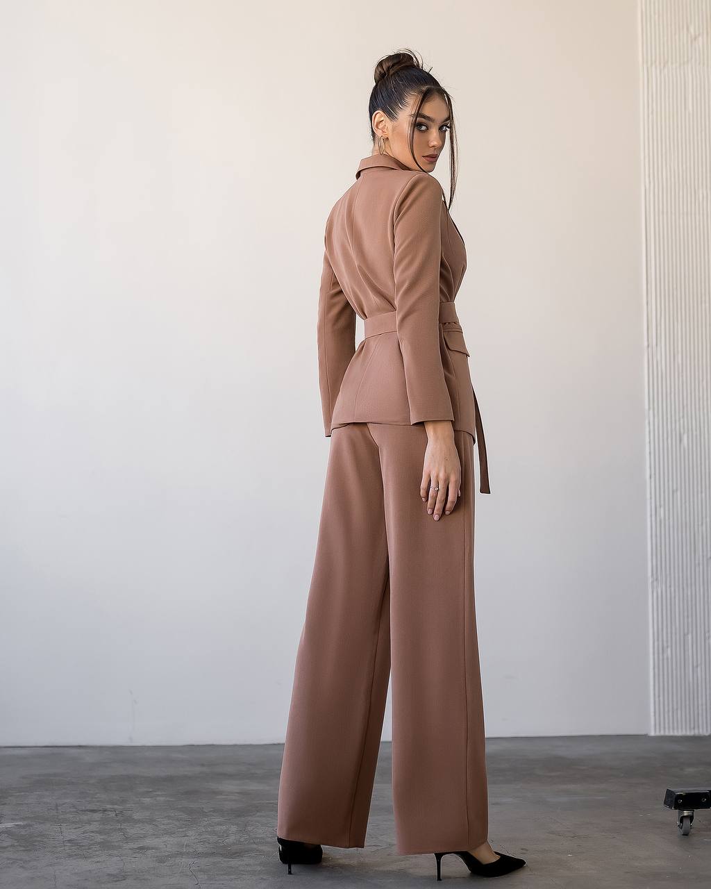 Woman wearing a brown suit standing against a plain wall.
