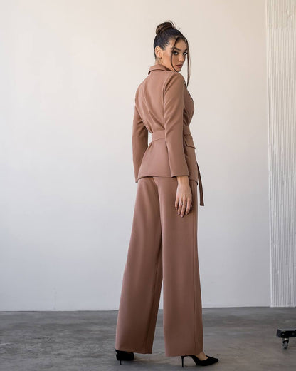 Woman wearing a brown suit standing against a plain wall.
