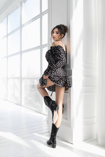 Woman in a black and white polka dot dress and black boots standing in a bright room with large windows.