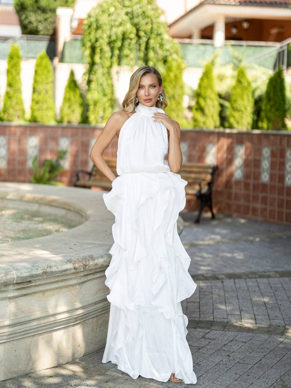 A woman wearing a white sleeveless halter-neck maxi dress with a high gathered neckline and dramatic cascading ruffles, standing outdoors in front of a brick wall and greenery.