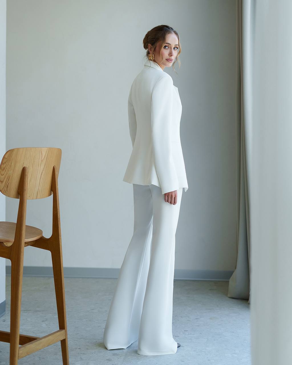 Woman in a white outfit standing in a minimalistic room with a wooden chair.