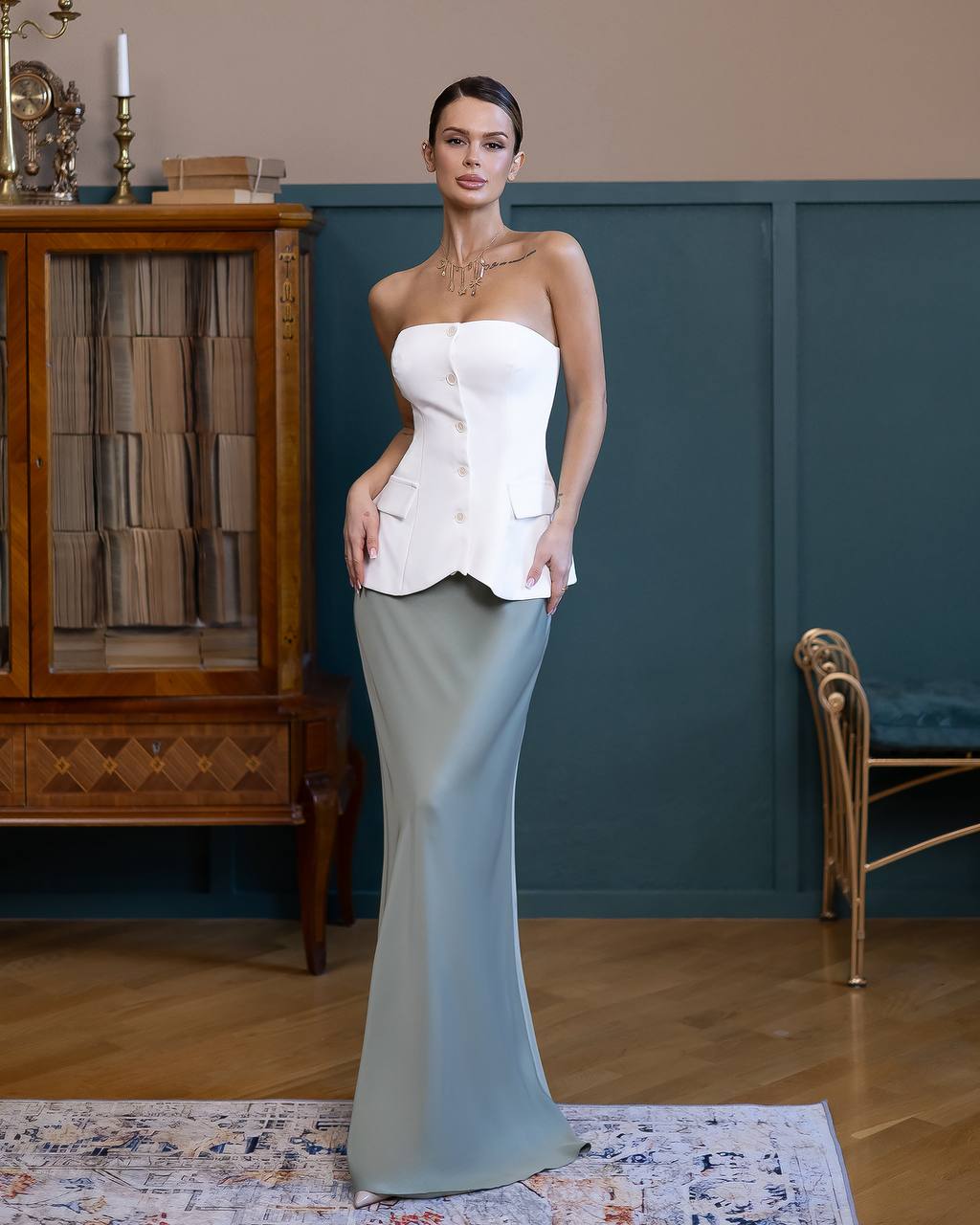 Woman in a strapless white top and long light blue skirt standing in a room with wooden furniture and a blue wall.