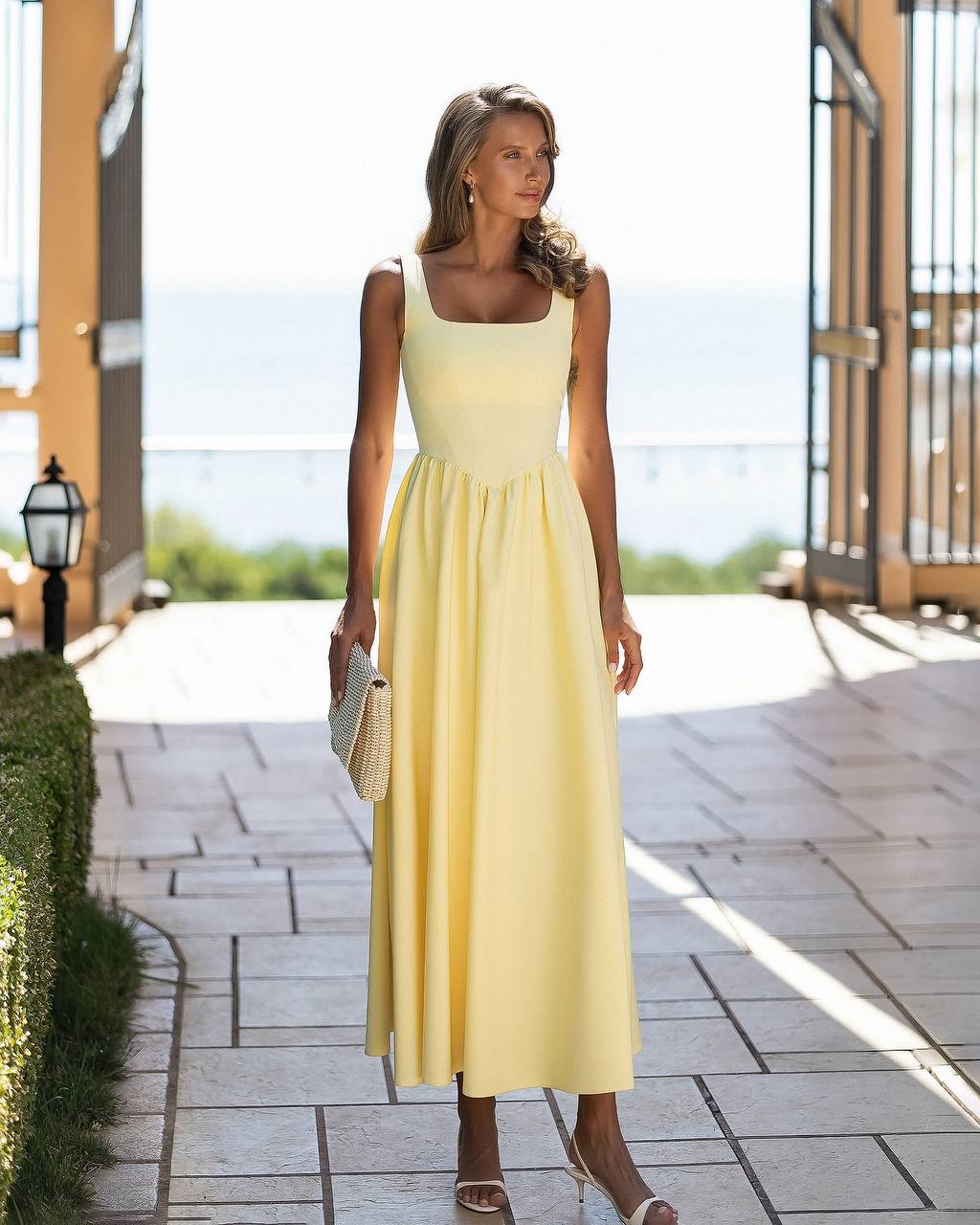 A woman wearing a pale yellow sleeveless midi dress with a square neckline and a gathered waist, standing on a stone walkway in a bright outdoor setting.
