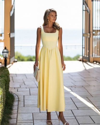 A woman wearing a pale yellow sleeveless midi dress with a square neckline and a gathered waist, standing on a stone walkway in a bright outdoor setting.
