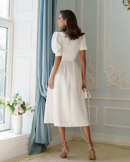 Woman in a white dress standing in a room with decorative walls and a window.