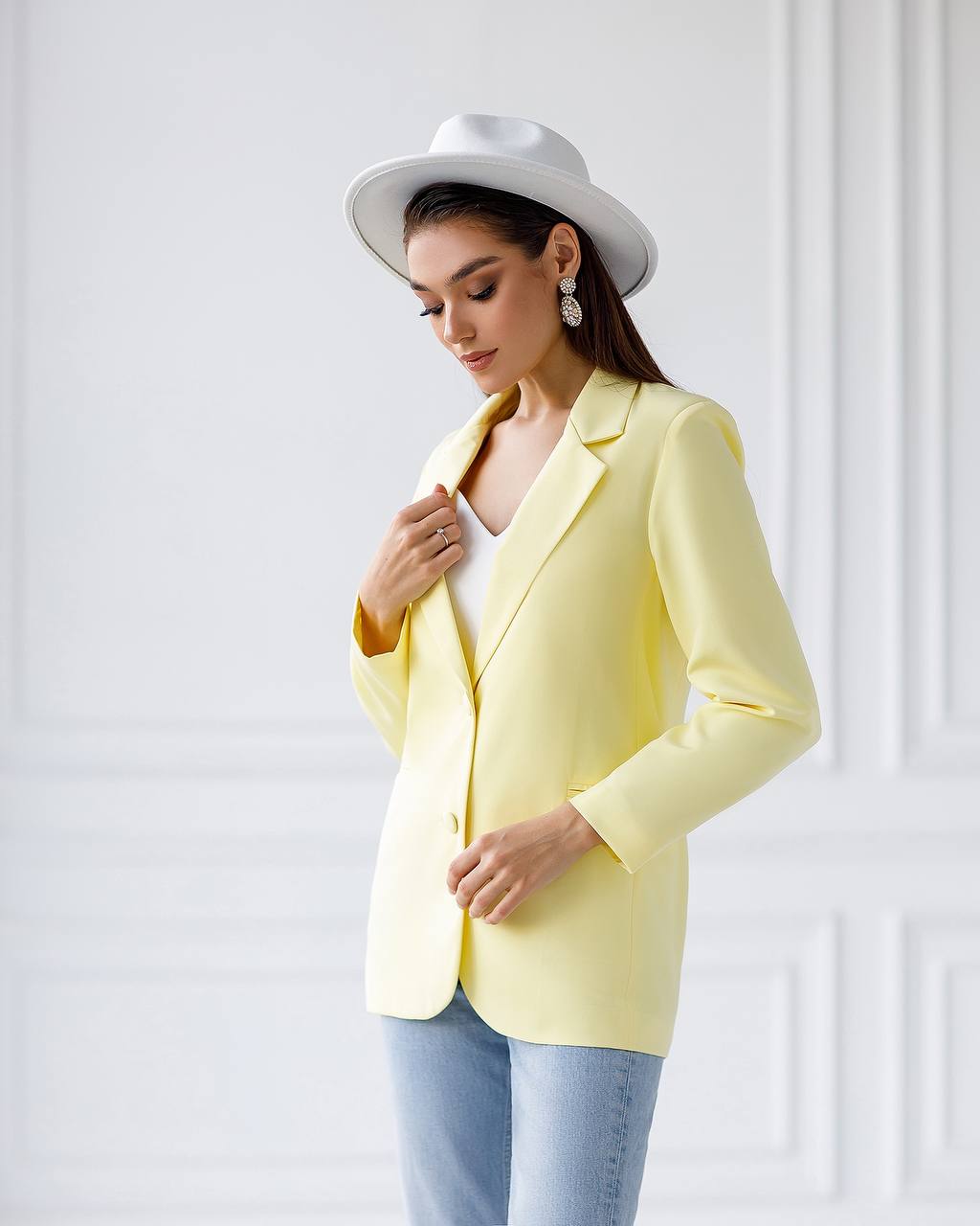 A woman wearing a pale yellow blazer, a white wide-brim hat, and light blue jeans, posing against a white paneled wall.