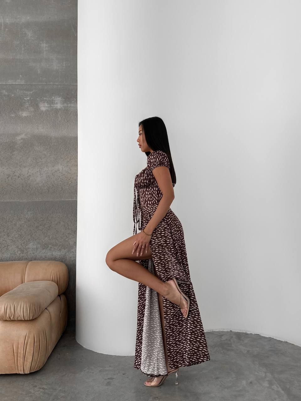 Woman in a patterned dress standing against a white wall with a beige sofa.