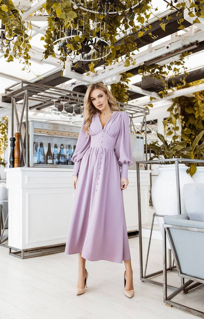 Woman in a lavender dress standing in an outdoor setting with plants and a bar.