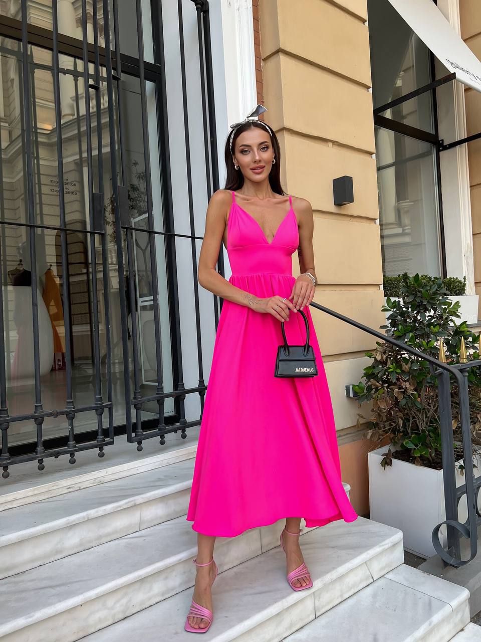 A woman stands on outdoor stone steps in front of a beige building, wearing a bright hot pink midi dress with thin straps and a gathered waist. She is holding a small black handbag and wearing pink heeled sandals.